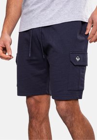 Homme portant un short cargo noir avec une taille à cordon de serrage et une poche plaquée sur la cuisse, assorti d'un t-shirt gris clair.