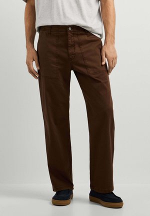 Hombre con pantalones rectos de color marrón oscuro con bolsillos parche y zapatos azul marino con suelas marrón claro sobre un fondo claro y liso.