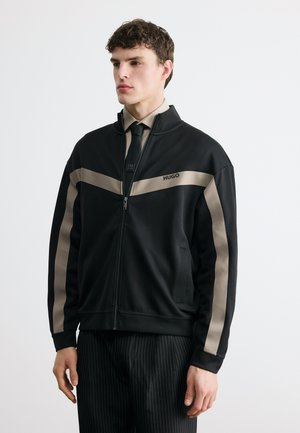 Mladý muž v čiernej bundě na zips s béžovými pruhmi a logom "HUGO", v kombinácii s béžovou košeľou, čiernou kravatou a čiernymi nohavicami s tenkými pásikmi.