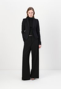 Crni oblikovani sako uparen s crnom turtleneck majicom i hlačama širokih nogavica. Glatka tkanina s preciznim krojem i minimalističkim dizajnerskim elementima.