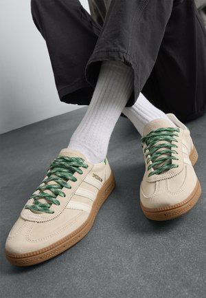 Zapatillas beige con cordones verdes y suelas de goma, llevadas con calcetines blancos de canalé y pantalones negros, con los pies cruzados sobre un suelo gris.