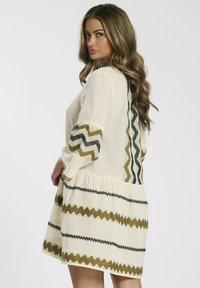 Robe beige clair avec des motifs en zigzag olive et noir. Dotée de manches longues, d'un ourlet arrondi et d'une coupe décontractée. Tissu texturé.