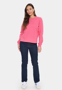Felpa rosa con scollo rotondo e maniche lunghe, abbinata a jeans blu scuro e sneakers multicolori. Tessuto morbido, vestibilità casual.