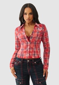 Czerwona koszula w kratę na guziki z długimi rękawami i dopasowanym krojem. Z białymi akcentami i kołnierzykiem, zestawiona z ciemnymi dżinsami z czerwonym przeszyciem.