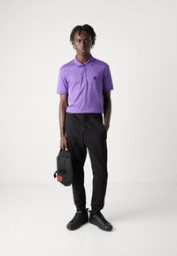 Lila polo skjorta med en logotyp, svarta joggers och svarta sneakers. Håller en liten svart väska med röd detalj. Enkelt design och passform.