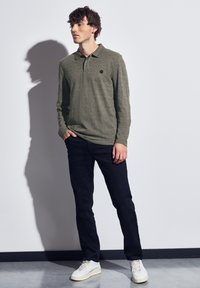 Polo olive à manches longues avec une texture subtile, jean noir et baskets blanches. Présente un petit logo rond sur la poitrine.