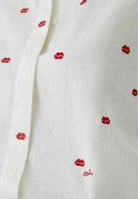White blouse featuring red embroidered lips scattered throughout, with the word "oui" embroidered in cursive near the bottom. Soft texture.