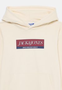 Sweat à capuche beige avec poche frontale arborant un logo rectangulaire rouge et bleu avec l'inscription "JACK & JONES SINCE NINETEEN NINETY."