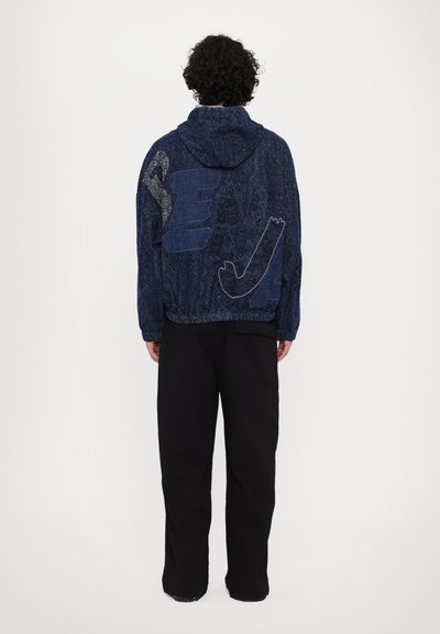 Veste à capuche en tissu bleu foncé avec des motifs graphiques contrastés au dos. La veste présente des poignets et un ourlet élastiques, associée à un pantalon noir.