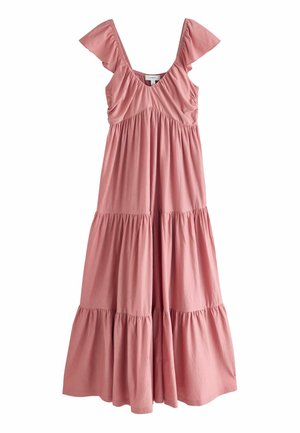 Kvinde i en pink lagdelt maxikjole, der står på en sandstrand med store klipper og havbølger i baggrunden.