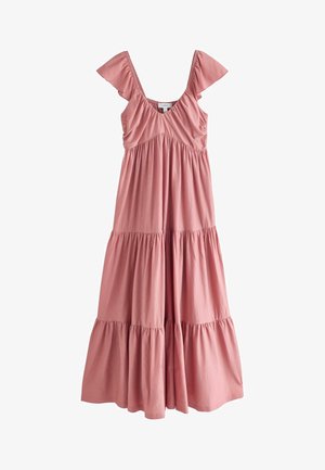 Naine roosas kihilise maksikleidi seljas seisab liivarannal, taga suured kivid ja ookeani lained.