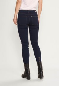 Skinny jeans in donkerblauwe denim, met een hoge taille, achterzakken en ritsen bij de enkels. Afgewerkt met oranje stiksels.