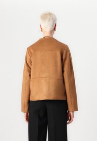 Bruine suède jas met een cropped ontwerp, met verlaagde schouders en een horizontale naad over de rug. Gecombineerd met zwarte broeken.
