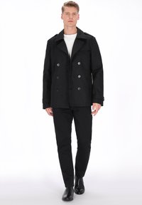 Sort dobbeltradet pea coat med store knapper og skulderflapper, båret over en hvid skjorte og sorte bukser, kombineret med sorte sko.