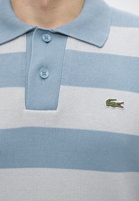 Gros plan d'un polo rayé bleu et blanc avec deux boutons fermés et un logo crocodile vert brodé sur la poitrine.