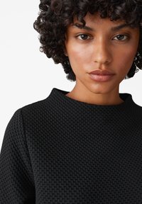 Top nero con texture e scollatura rotonda, caratterizzato da un motivo intrecciato. Pelle liscia visibile, con acconciatura riccia che incornicia il viso.