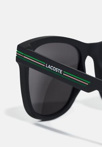 Ochelari de soare negri cu o finisare mată, având un logo pe braț și dungi de accent verzi și roz de-a lungul ramei. Lentile întunecate, nuanțate.