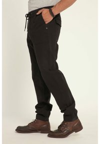 Pantaloni cargo neri con vita elastica, cordoncino, multiple tasche e polsini affusolati. Indossati con stivali marroni con lacci.