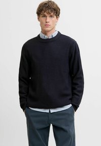 Suéter de punto waffle en color navy con cuello redondo, combinado con una camisa de rayas azul claro y blanca. Presenta puños y dobladillo acanalados. Pantalones oscuros.