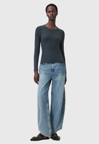 Top a maniche lunghe verde scuro abbinato a jeans larghi di un azzurro chiaro. I jeans presentano un orlo sfilacciato, e le scarpe sono sandali piatti neri.