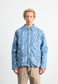Chaqueta de mezclilla azul acolchada con un patrón abstracto blanco, cuello puntiagudo y cierre de botones, con cuerdas ajustables en el dobladillo.
