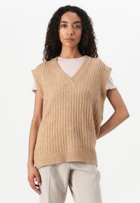 Beige ribbet strikket vest med V-hals, korte ermer og en teksturert overflate. Bært over en lys rosa topp, sammen med lysfargede bukser.