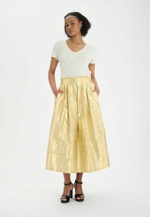 Jupe maxi plissée dorée avec une taille élastique, dotée de poches, associée à un t-shirt blanc à col en V et des sandales à talons noirs.