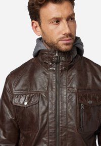 Braune Lederjacke mit hohem Kragen, grauer Hoodie-Fütterung, mehreren Taschen und metallenen Reißverschlussdetails; strukturierte Oberfläche und durchdachtes Design.