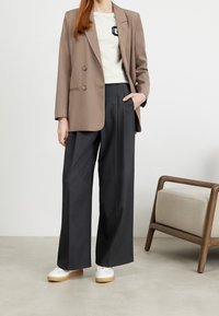 Braun doppelt knöpfbare Blazer, graue weit geschnittene Hose, weiße Sneakers mit einer hellbraunen Sohle und ein helles Oberteil mit einem Aufnäher.