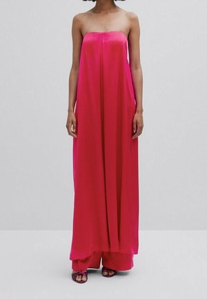 Robe longue rose vif sans bretelles avec larges jambes portée avec des sandales foncées sur fond uni.