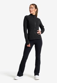 Svart zip-jacka med figursydd design, hög krage och elastiskt tyg. I kombination med svarta byxor och vita sportskor. Slen struktur.