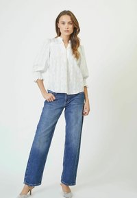 Blusa bianca con un sottile motivo testurizzato, maniche lunghe a sbuffo e colletto alla cinese. Abbinata a jeans in denim blu a gamba larga e tacchi trasparenti.