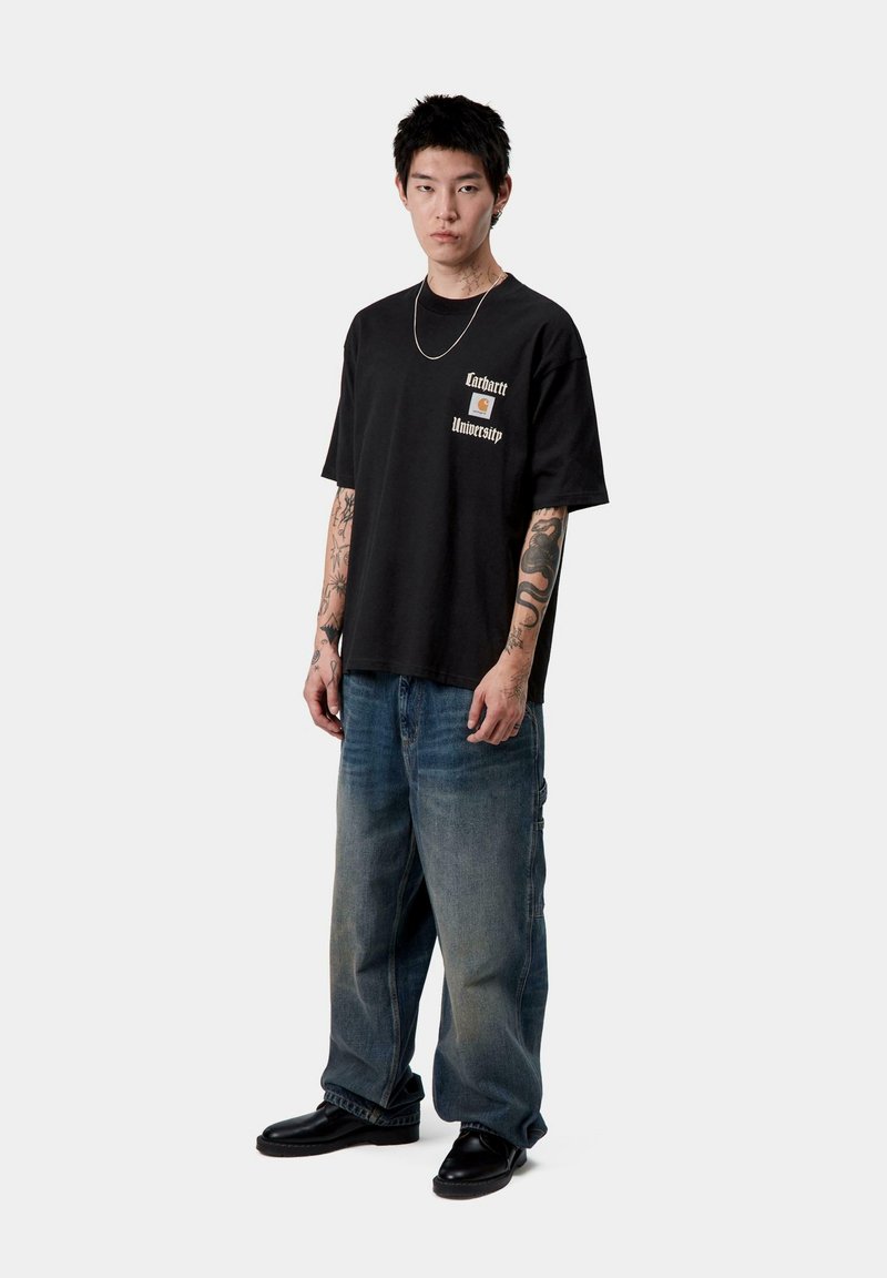 T-shirt nera in cotone con logo stampato sul lato sinistro del petto, abbinata a jeans baggy blu chiaro e scarpe nere. Il modello ha dei tatuaggi e capelli corti.