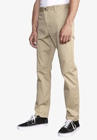 Beige cotton chinos with a straight-leg fit, front pockets, and a zipper fly. Paired with black sneakers featuring white laces and soles.