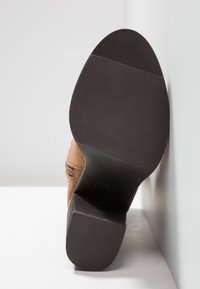 Semelle d'une botte en daim brun avec un talon en caoutchouc noir et une semelle rainurée, inclinée contre un fond blanc de mur et de sol.
