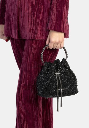 Borsa a secchiello nera con superficie testurizzata, impreziosita da borchie e chiusura con coulisse, tenuta da una persona che indossa un abito bordeaux in velluto.