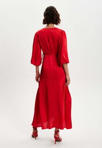 Red, flowy maxi dress with puffed sleeves, belted waist, and ankle-length hem. Smooth texture with a lightweight fabric. Stiletto heels in red.