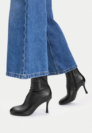 Beine in weiten blauen Jeans und schwarzen Leder-Stiefeletten mit hohen Absätzen und seitlichen Reißverschlüssen vor weißem Hintergrund.