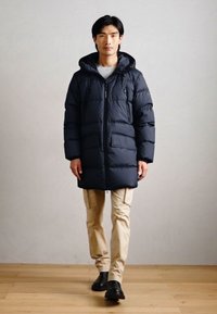 Férfi fekete kapucnis puffermellényben, bézs cargo nadrágban és fekete cipőben, egy sima világosszürke fal előtt áll.