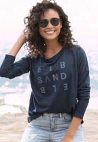 Navy long-sleeve shirt with oversized fit, featuring gray upside-down text design; paired with light-wash distressed jeans.