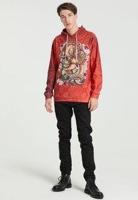 Rød hættetrøje med et farverigt dragegrafik, sammen med en tekstureret baggrund og hvide snore. Saty med sorte slim-fit bukser og støvler.