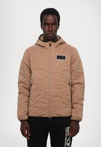 Quiltet beige jakke med hætte, lynlåslukning og et sort logo-patch. Har elastiske manchetter og en glat tekstur.
