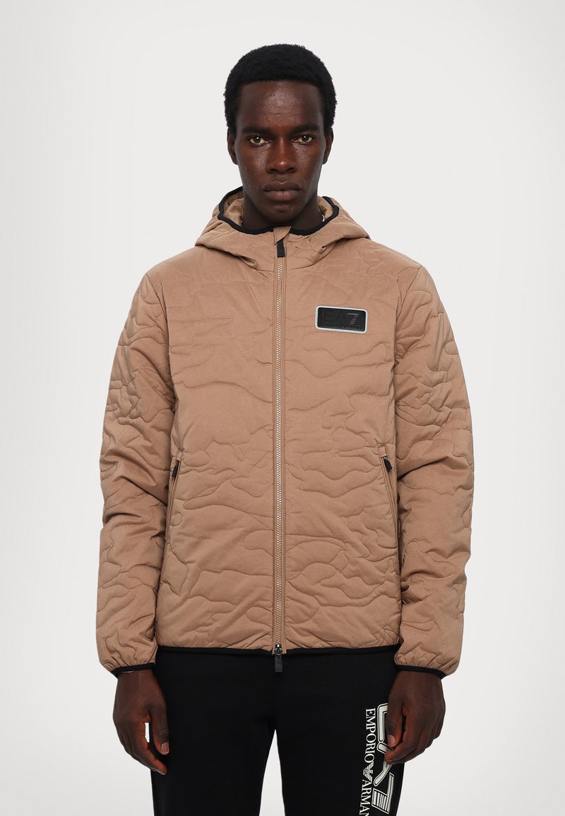 Quiltet beige jakke med hætte, lynlåslukning og et sort logo-patch. Har elastiske manchetter og en glat tekstur.