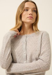 Cardigan en tricot gris clair avec un motif en diamant, doté d'un col rond, de manches longues et de cinq boutons à l'avant avec une finition texturée.