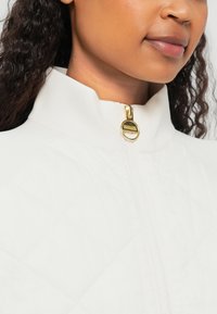 Casaco branco acolchoado com gola alta e detalhe de fecho em dourado. Close-up mostrando a textura e os elementos de design.