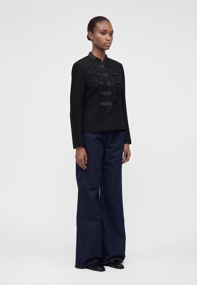 Femme debout portant une veste noire boutonnée avec des détails en satin, un pantalon large bleu foncé et des chaussures noires devant un fond clair uni.