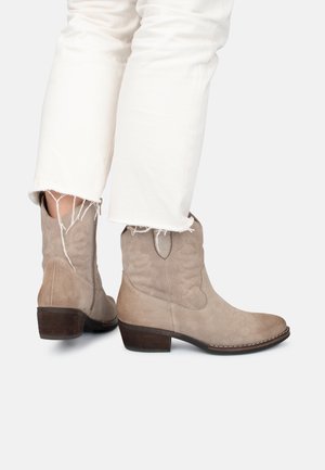 Stivali ankle beige in suede con tacco basso a blocco, caratterizzati da cuciture decorative sui lati e punta arrotondata. Indossati con jeans bianchi sfilacciati.