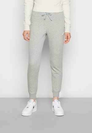 Personne portant un pantalon de jogging Nike gris clair avec une taille à cordon et des baskets blanches Nike, debout devant un fond uni.
