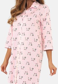 Kobieta w różowej i białej koszulowej sukience w paski z ilustracjami małych psów, z rękawami do trzech czwarte, zapinana na guziki.
