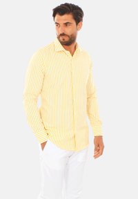 Camicia a righe gialle e bianche con maniche lunghe, vestibilità su misura e colletto. Il modello la indossa con pantaloni bianchi, mani in tasca.